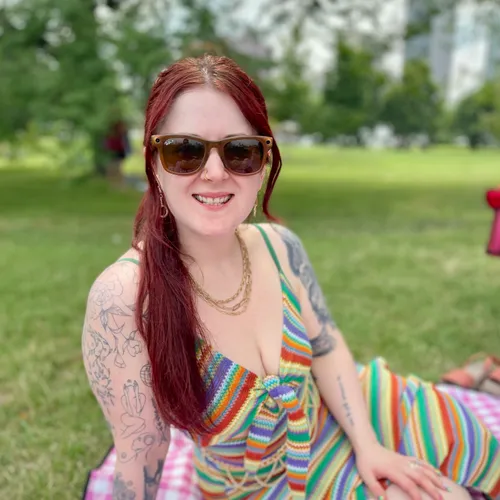 Tiana McCrory smiling widely outdoors in a park, wearing sunglasses and a colorful striped top, seated on a picnic blanket with green grass and trees behind her.