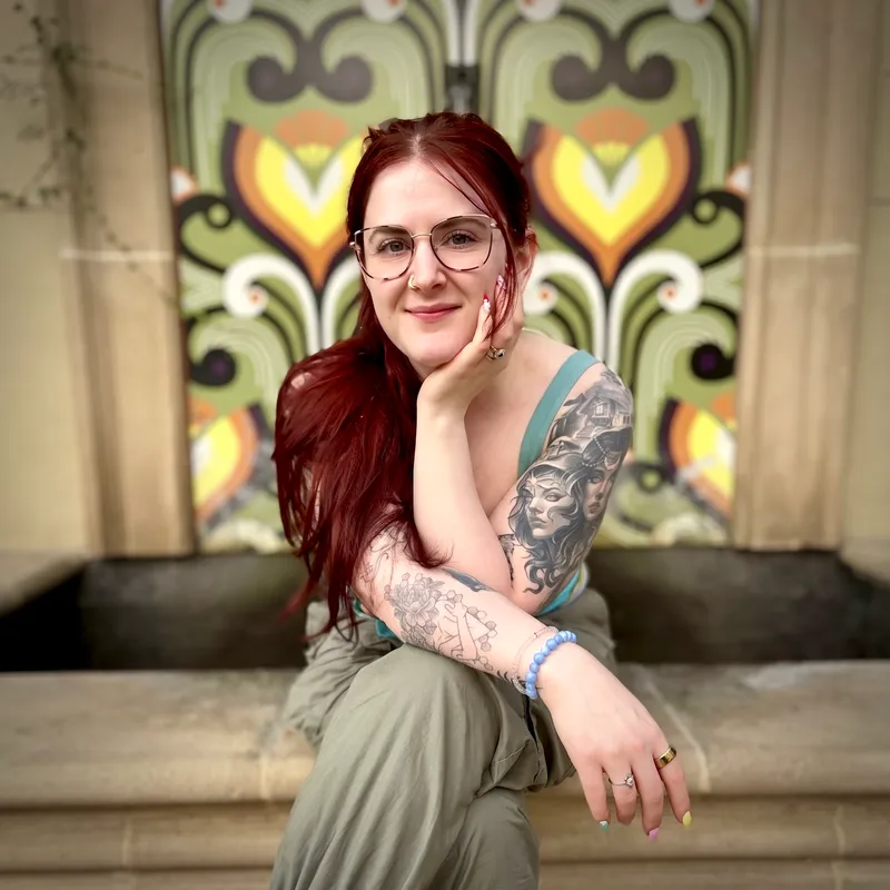 Tiana McCrory smiling gently with her chin resting on her hand, seated in front of a colorful ornate floral mural. She has long red hair, glasses, and visible tattoo artwork on her arm.